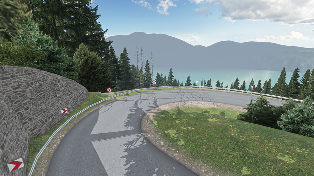 A scenic mountain road with a sharp hairpin turn, bordered by a stone wall on one side and dense evergreen trees on the other. The road overlooks a misty lake in the distance, surrounded by towering mountains under a partly cloudy sky.