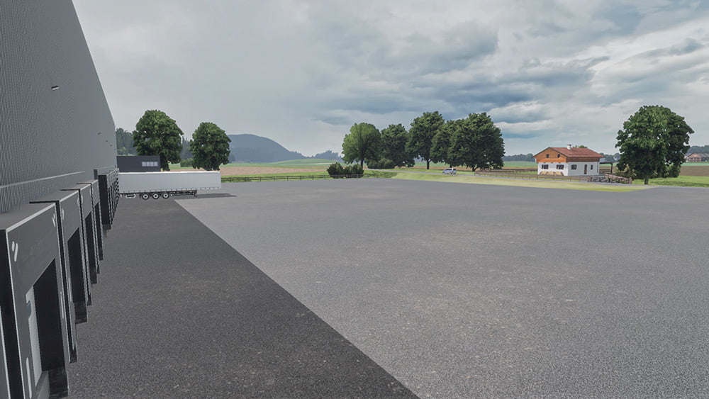 A wide open truck parking lot with loading docks attached to a large warehouse. A semi-truck trailer is positioned near the building, and in the background, there are trees and a small country house with a green field stretching towards distant hills under a cloudy sky.