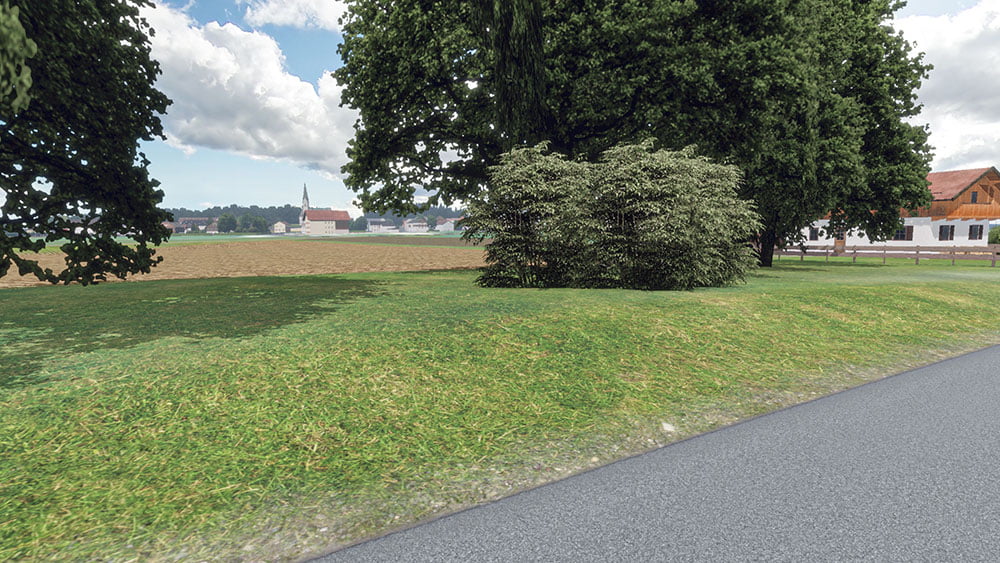 A serene countryside view featuring a green patch of grass bordered by trees and bushes. A paved road runs alongside the scene, and a distant village with a church tower is visible across the fields under a partly cloudy sky.
